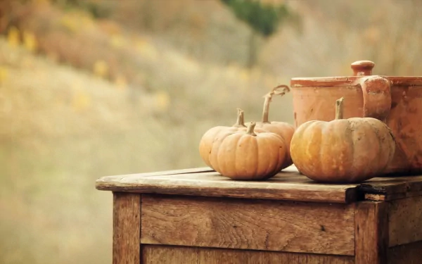 HD desktop wallpaper featuring two pumpkins and rustic kitchenware on a wooden table with a blurred autumnal background.