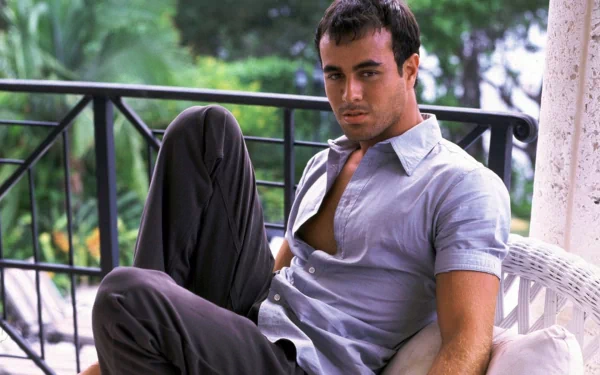 A high-definition desktop wallpaper featuring Enrique Iglesias, relaxed and stylishly dressed, seated outdoors with lush greenery in the background.