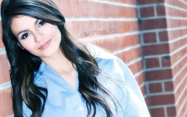 A high-definition desktop wallpaper featuring Victoria Justice, smiling softly against a textured brick wall, showcasing her long, flowing hair and casual style.