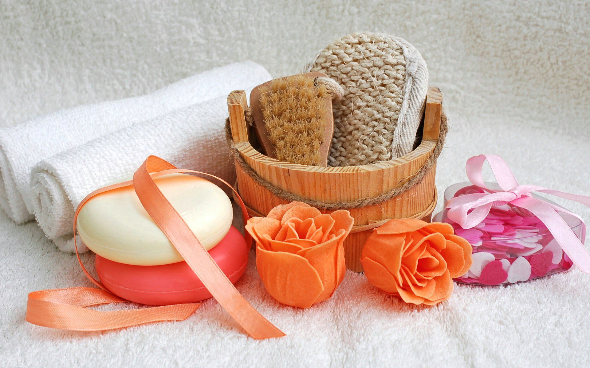 Man-made spa arrangement with soaps, a wooden bucket holding exfoliating tools, orange rose-shaped candles, and rolled white towels on a soft textured surface.