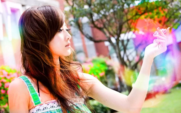A vibrant HD wallpaper featuring Mikako Zhang Kaijie, a woman with long hair in a colorful outdoor setting, looking intently at a bright object in her hand.
