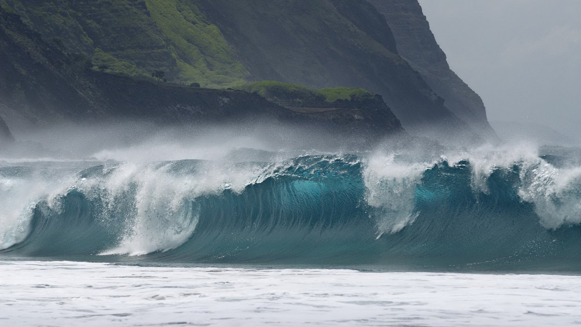 HD desktop wallpaper showcasing a powerful ocean wave crashing near a rocky, green mountainside under a misty sky, highlighting the beauty of nature.