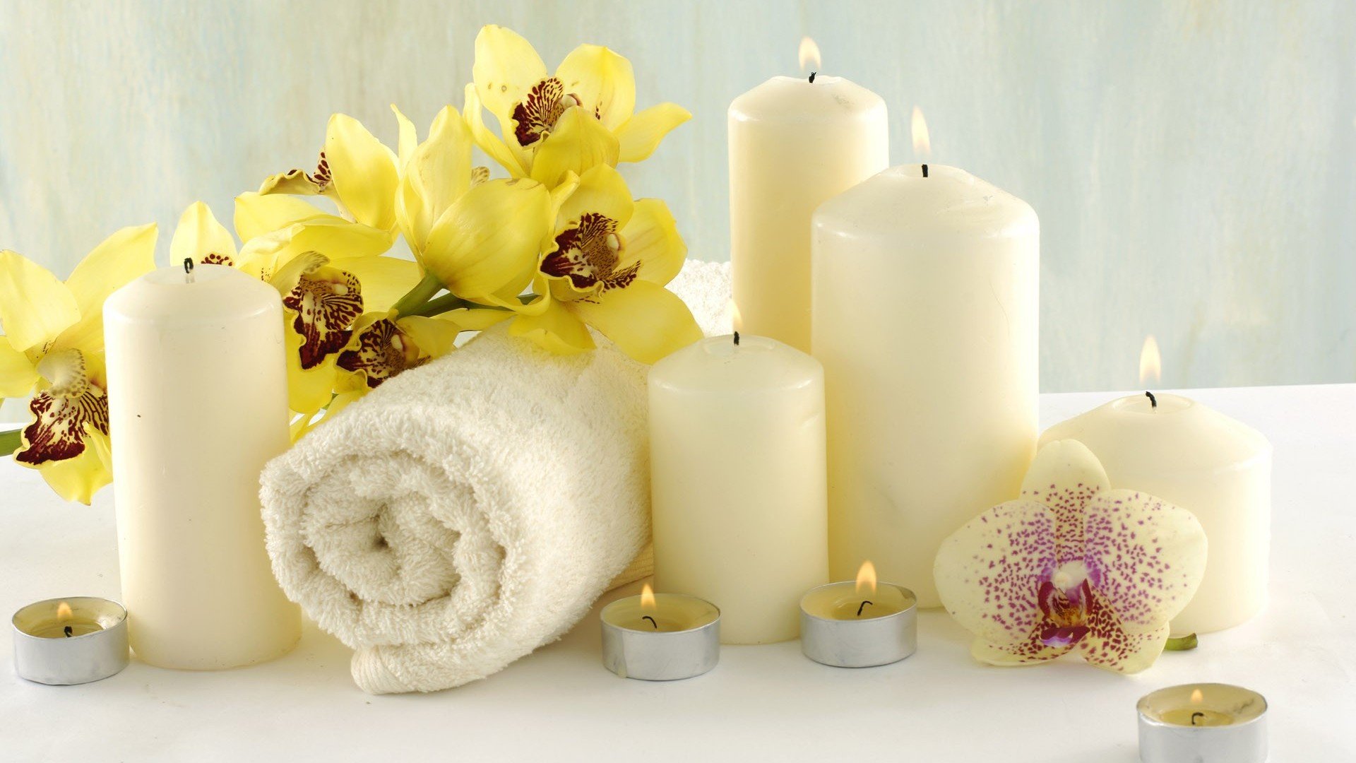 Still life HD wallpaper featuring yellow orchids, lit white candles, and a rolled white towel creating a serene spa atmosphere.
