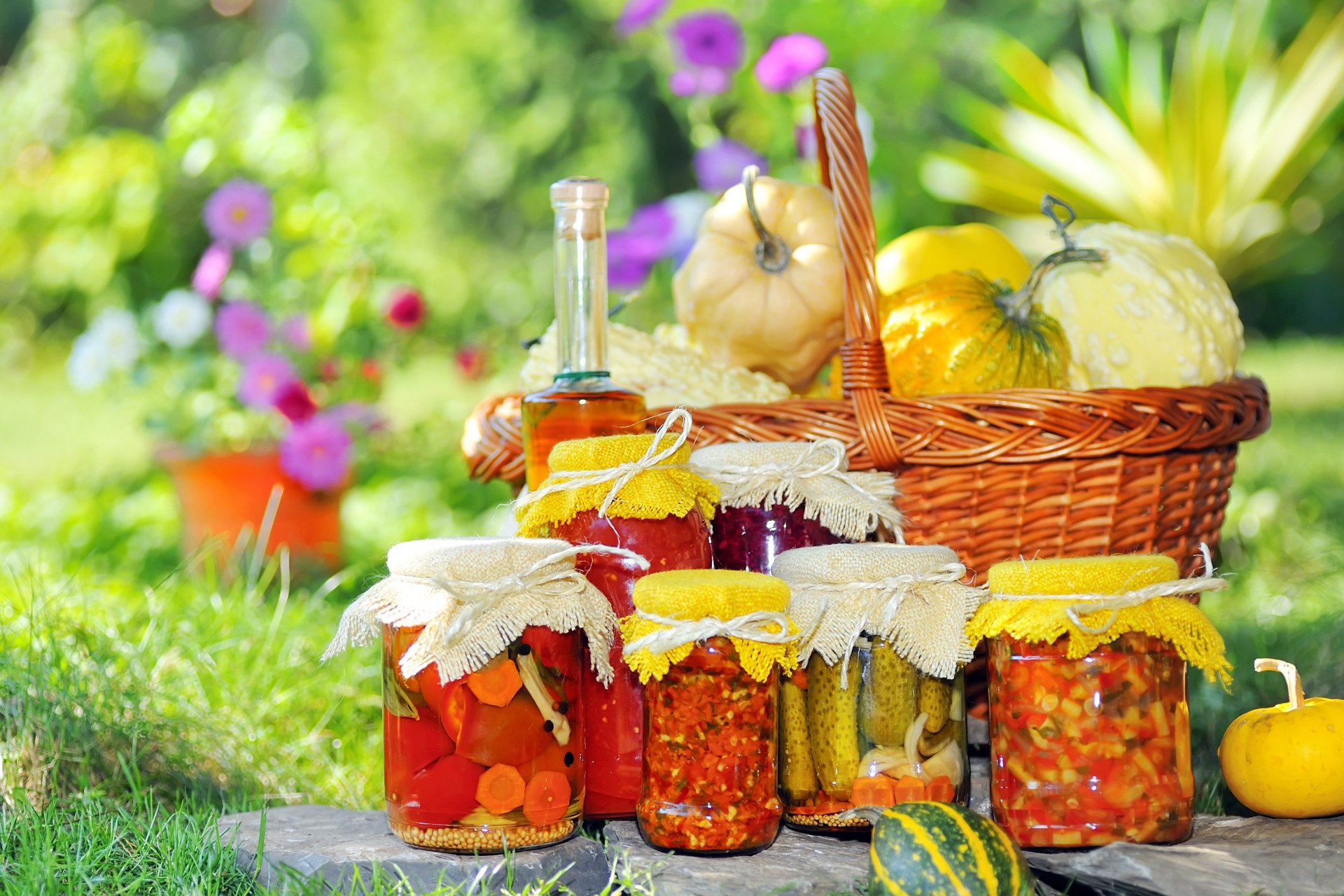 HD PC desktop wallpaper featuring a still life of assorted homemade preserved vegetables in jars and a basket of fresh gourds set outdoors on green grass with vibrant flowers.