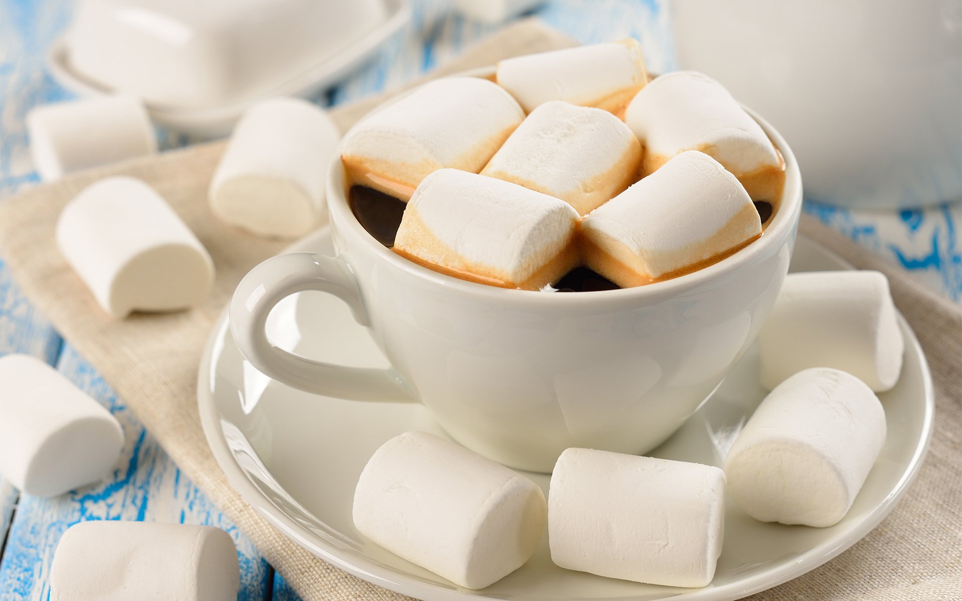 HD PC desktop wallpaper featuring a white cup filled with coffee topped with marshmallows, surrounded by scattered marshmallows on a rustic blue surface.