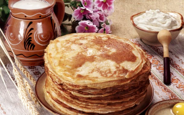 HD PC desktop wallpaper featuring a stack of golden crêpes accompanied by a rustic jug, bowl of cream, and pink flowers on a textured wooden surface.