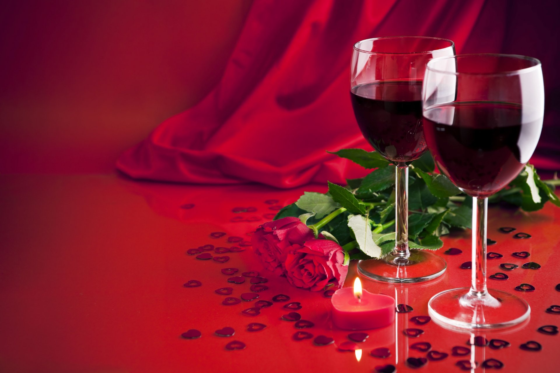Two glasses of red wine sit beside a rose and a flickering candle on a red backdrop, creating a romantic Valentine's Day scene in this vibrant 4K Ultra HD wallpaper.
