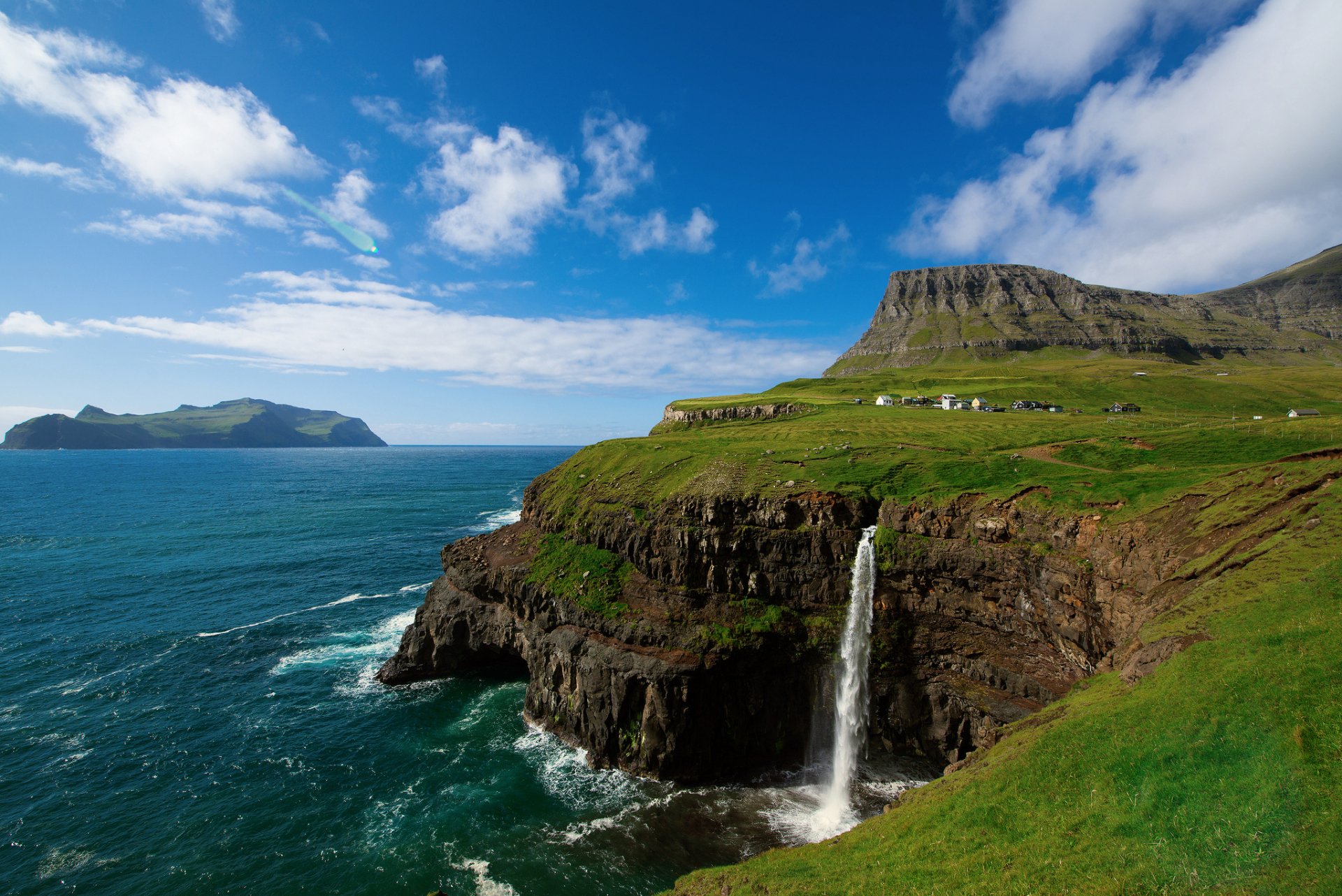 HD desktop wallpaper showcasing a vibrant natural landscape with a waterfall cascading off a green cliff into the ocean under a bright blue sky with scattered clouds.