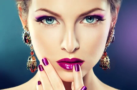 HD close-up of a woman with blue eyes, wearing bold purple lipstick and extravagant earrings. Her makeup is prominent, and she touches her face with matching purple nail polish. Elegant jewelry adorns her ears.