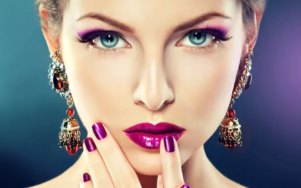 HD close-up of a woman with blue eyes, wearing bold purple lipstick and extravagant earrings. Her makeup is prominent, and she touches her face with matching purple nail polish. Elegant jewelry adorns her ears.