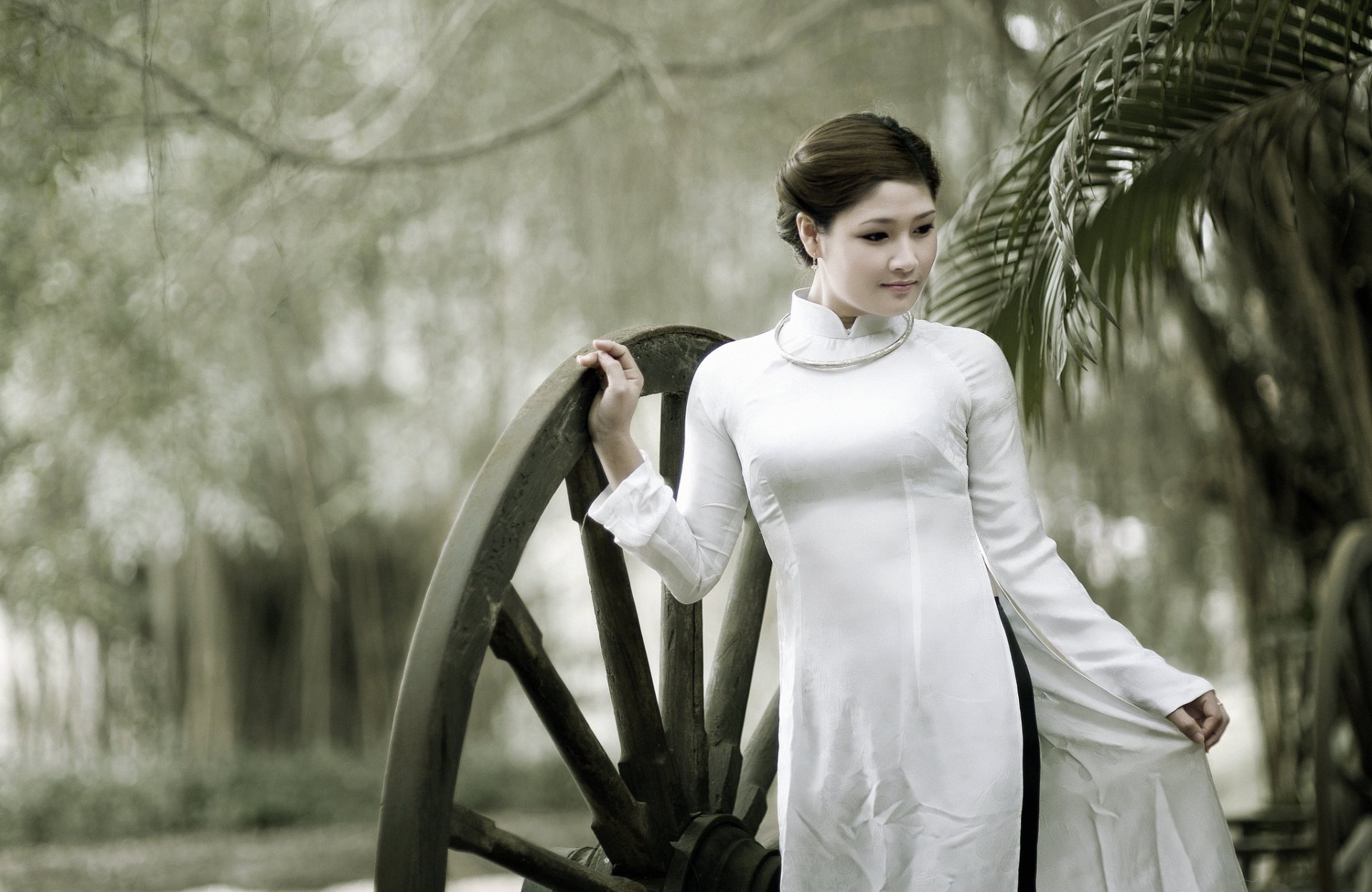 A serene portrait of an Asian woman in traditional attire, standing beside a large wooden wheel, set against a backdrop of blurred greenery. HD desktop wallpaper.