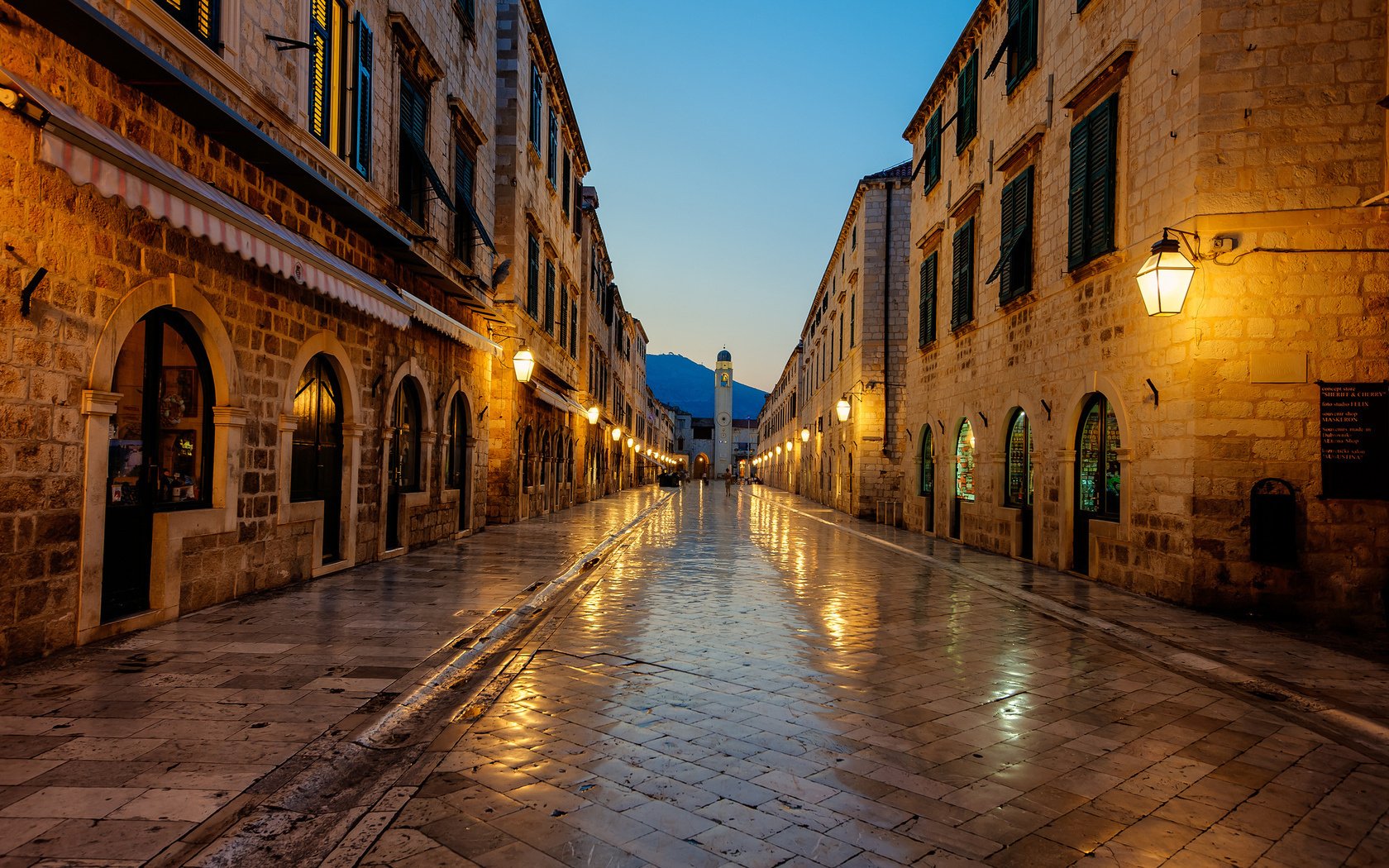 HD desktop wallpaper showcasing a man-made street scene in Dubrovnik with historic stone buildings and warm streetlights reflecting on wet pavement at dusk.