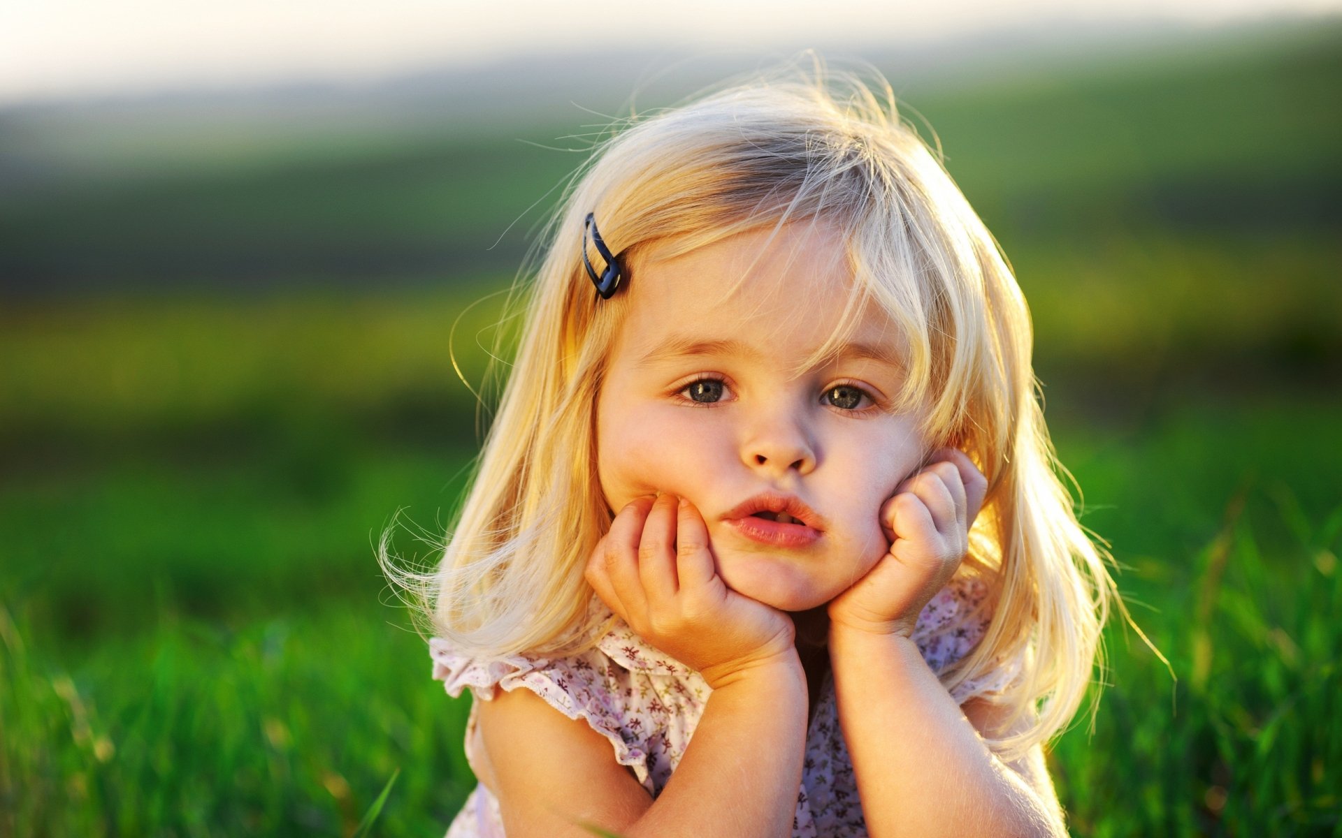 A cute blonde little girl rests her chin on her hands, gazing thoughtfully into the distance, surrounded by a serene green background. A charming photography piece for desktop wallpaper.