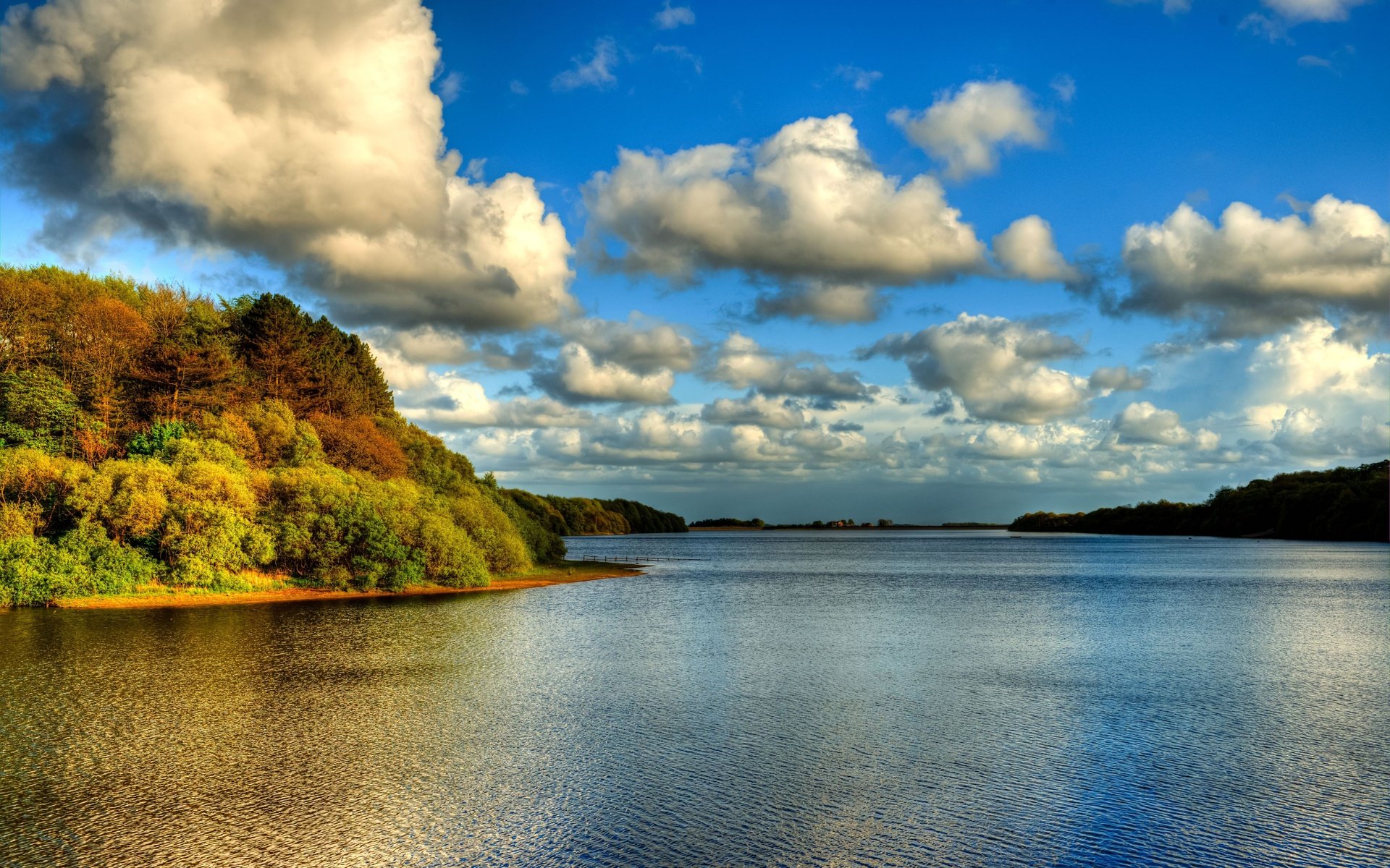 HD desktop wallpaper showcasing a tranquil lake surrounded by lush trees under a vibrant blue sky filled with fluffy clouds.