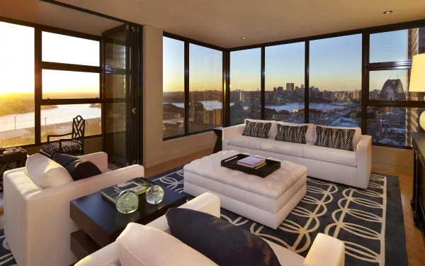 A modern, man-made room with large windows overlooking a city skyline at sunset, featuring white sofas and a patterned rug, captured in 4K Ultra HD for a PC desktop wallpaper.