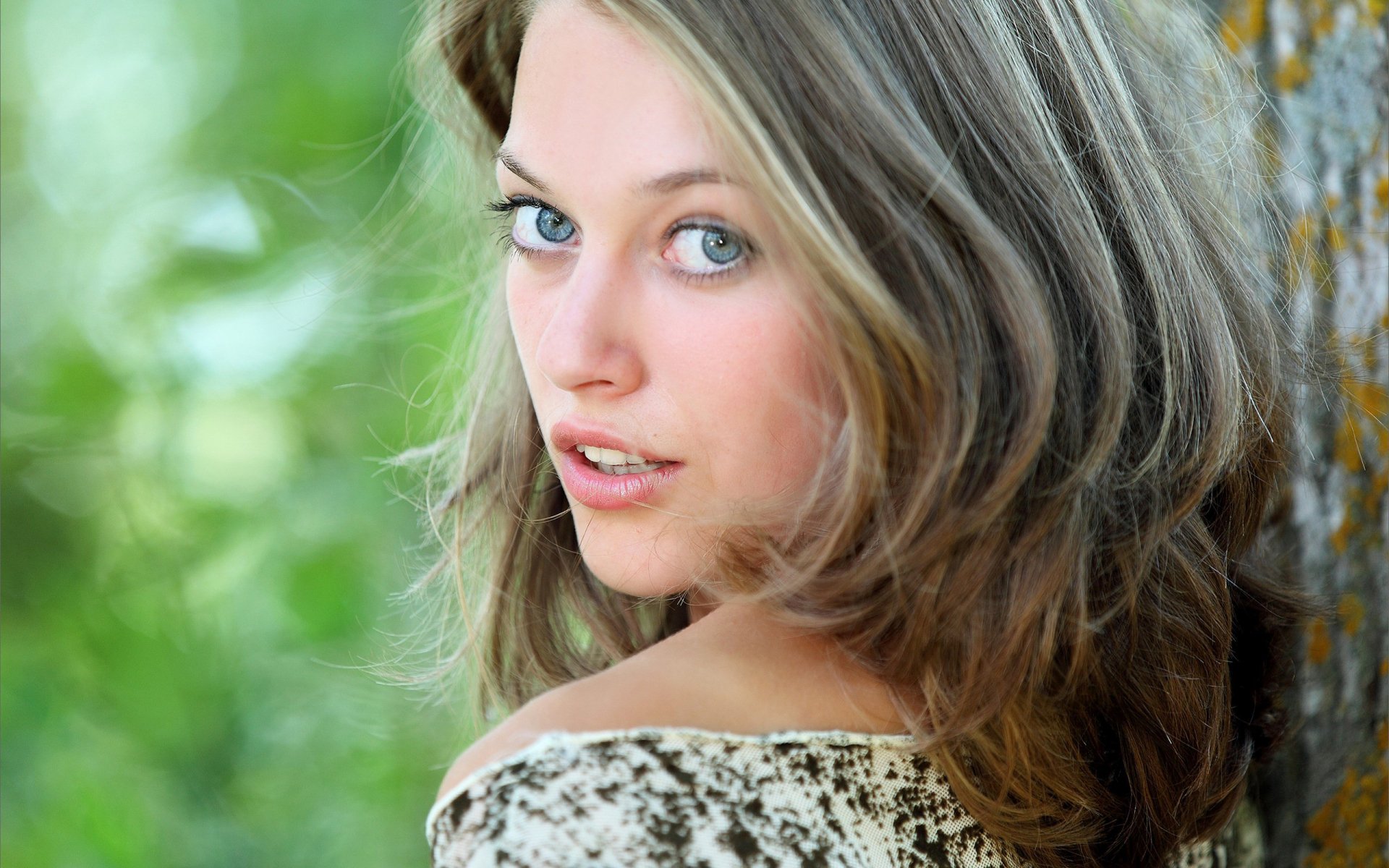 Close-up of a woman model leaning against a tree with soft-focus greenery, 2K Quad HD PC desktop wallpaper background.