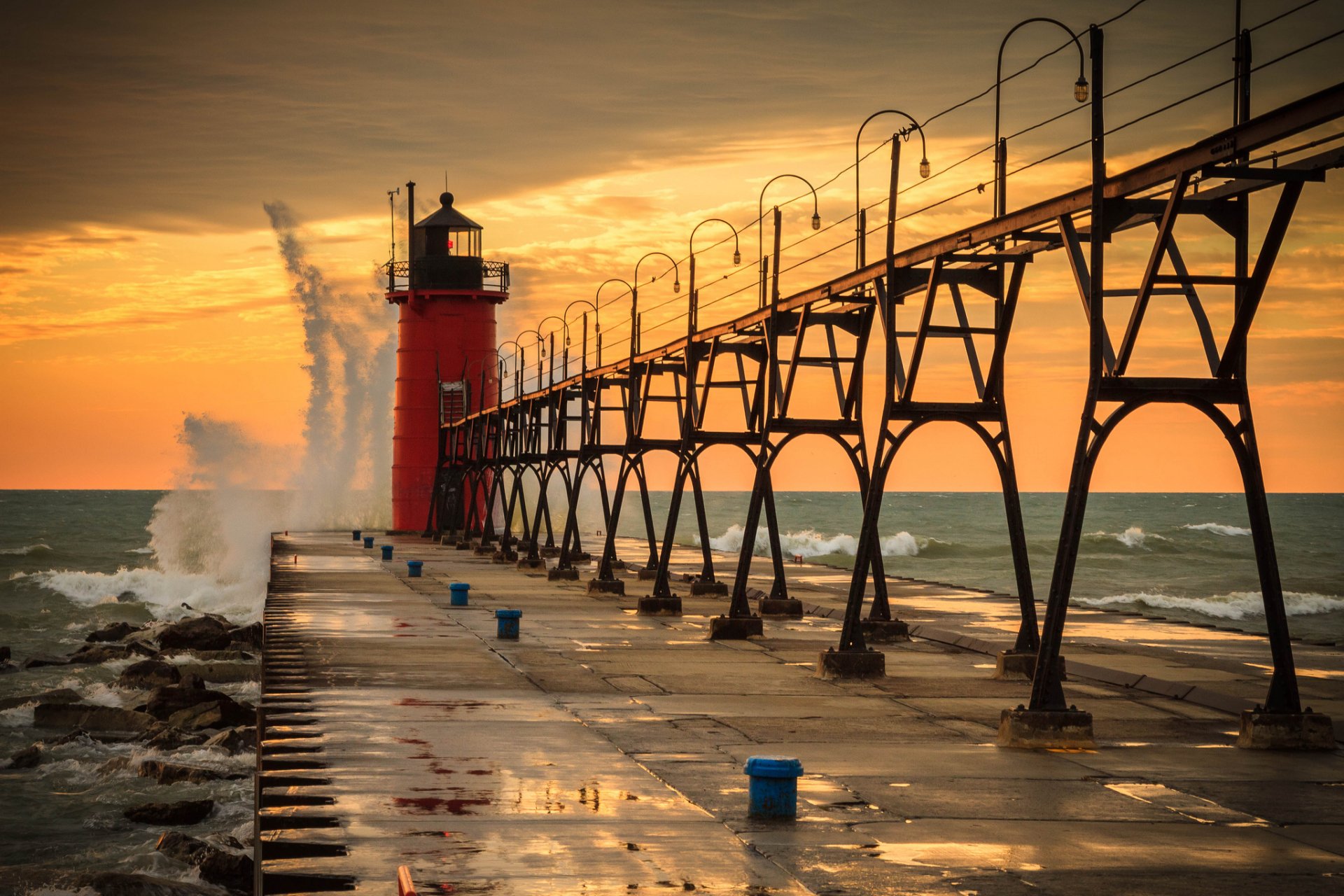 HD desktop wallpaper of a man-made lighthouse at sunset with waves crashing against the pier, featuring vibrant orange and yellow skies over a calm sea.