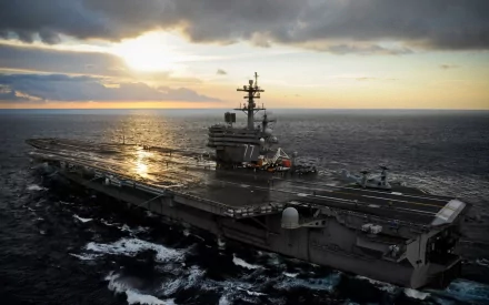 An aerial view of the USS George H.W. Bush (CVN-77), a military aircraft carrier, sailing through the ocean under a dramatic sunset backdrop.