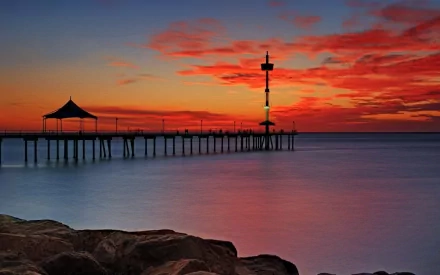 HD PC desktop wallpaper: man-made pier and tower silhouetted against a vivid red-orange sunset over calm sea, rocky foreground.