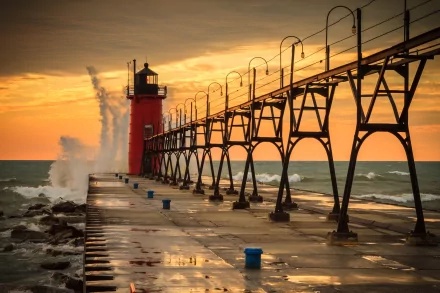 HD desktop wallpaper of a man-made lighthouse at sunset with waves crashing against the pier, featuring vibrant orange and yellow skies over a calm sea.