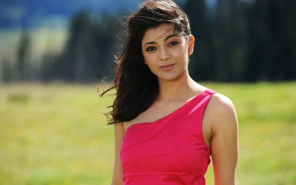 An HD desktop wallpaper featuring Bollywood actress Kajal Aggarwal standing outdoors in a pink dress, with a serene natural background.