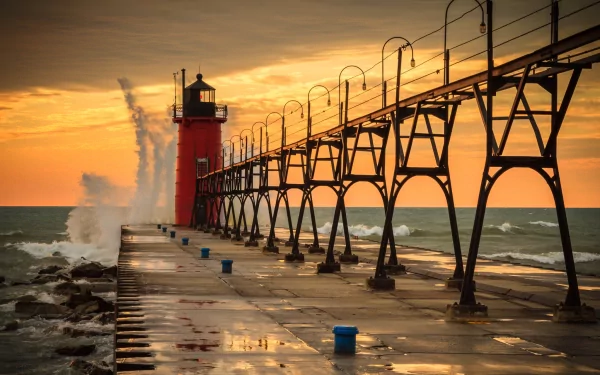 HD desktop wallpaper of a man-made lighthouse at sunset with waves crashing against the pier, featuring vibrant orange and yellow skies over a calm sea.