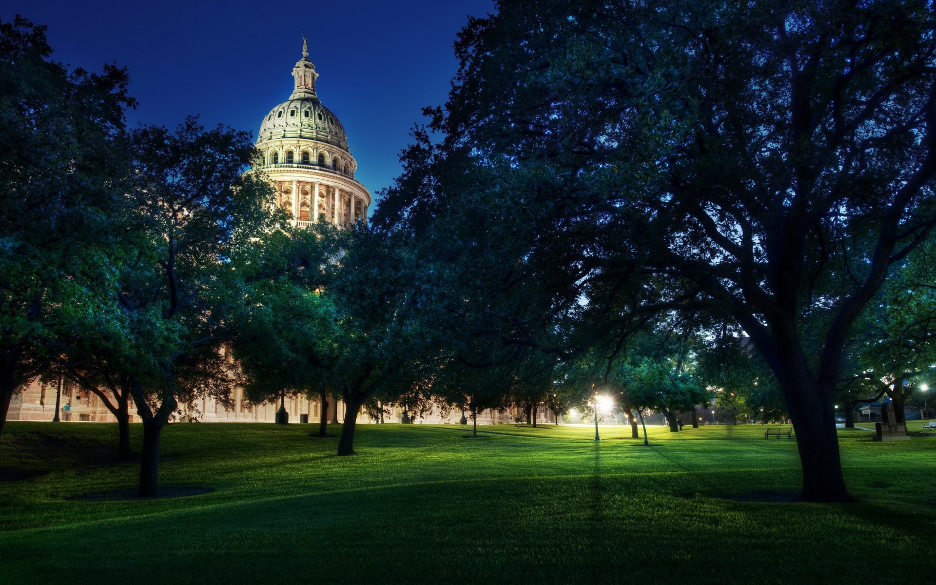 HD desktop wallpaper showing the illuminated dome of a historic capitol building at dusk, framed by trees on a lush green lawn, representing a man-made landmark.