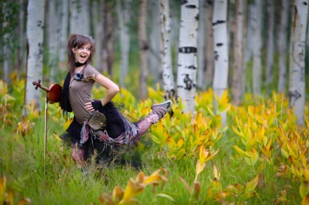 Lindsey Stirling joyfully poses with a violin in a vibrant, natural setting filled with tall trees and lush yellow foliage, capturing the spirit of music and nature in this stunning 4K Ultra HD wallpaper.