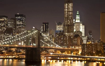 A stunning night view of Manhattan, featuring the iconic Brooklyn Bridge illuminated against a backdrop of city skyscrapers and shimmering waters. HD desktop wallpaper.