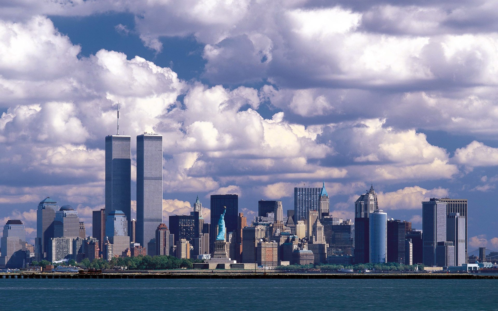 HD PC desktop wallpaper of Manhattan, featuring the Statue of Liberty and Twin Towers rising above New York's man-made skyline under a dramatic cloud-filled sky.