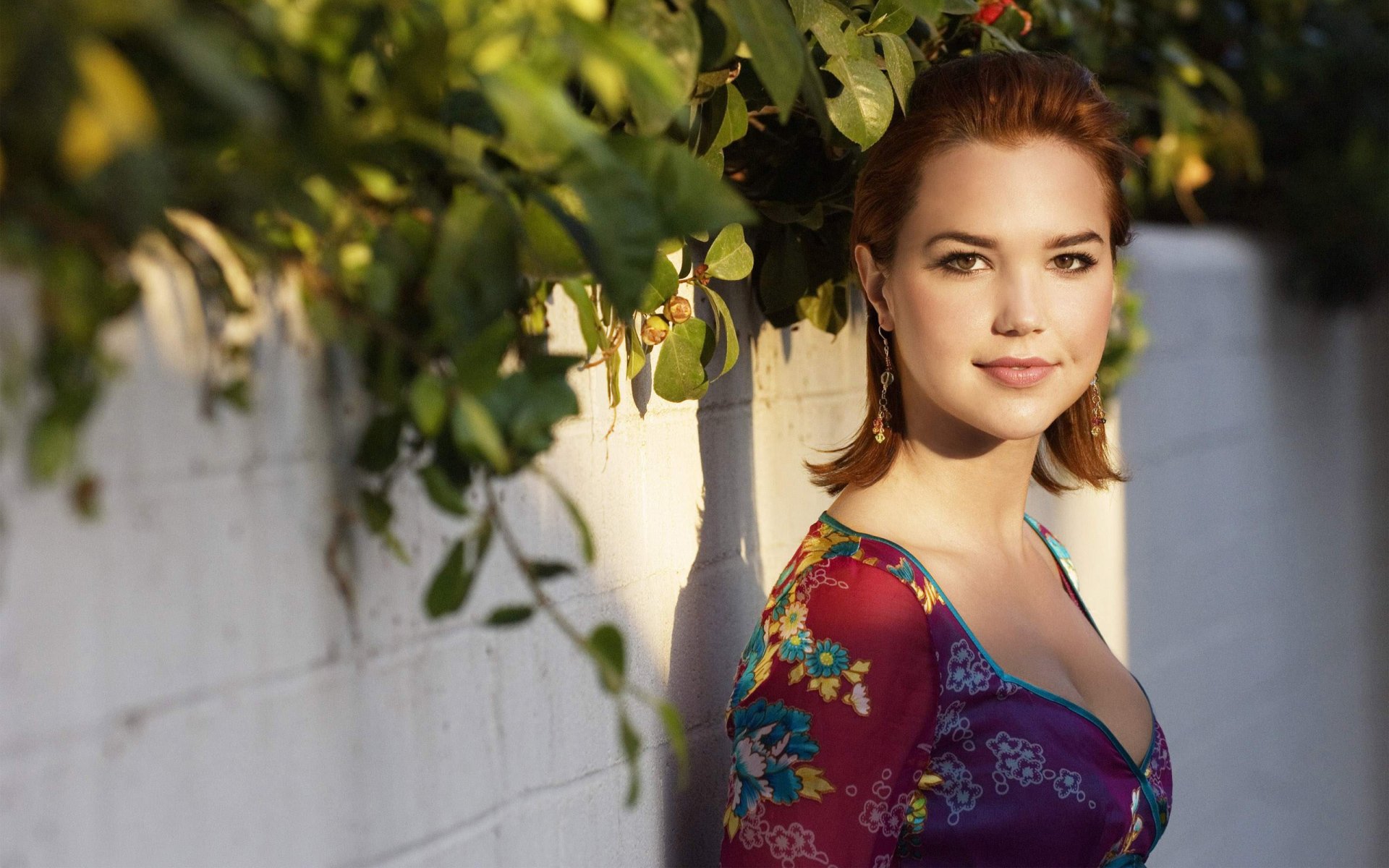 Sunlit portrait of a woman in a floral dress leaning against a white wall under leafy branches — 2K Quad HD PC desktop wallpaper/background.