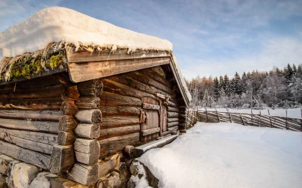HD PC desktop wallpaper: snow-covered man-made log cabin beside a split-rail fence, frozen clearing and frosted pine forest beneath a pale blue winter sky.