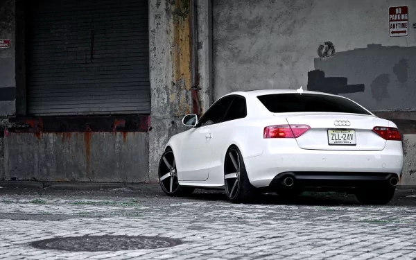 White Audi A5 parked by a weathered industrial wall on cobblestones, styled as an HD PC desktop wallpaper and background.