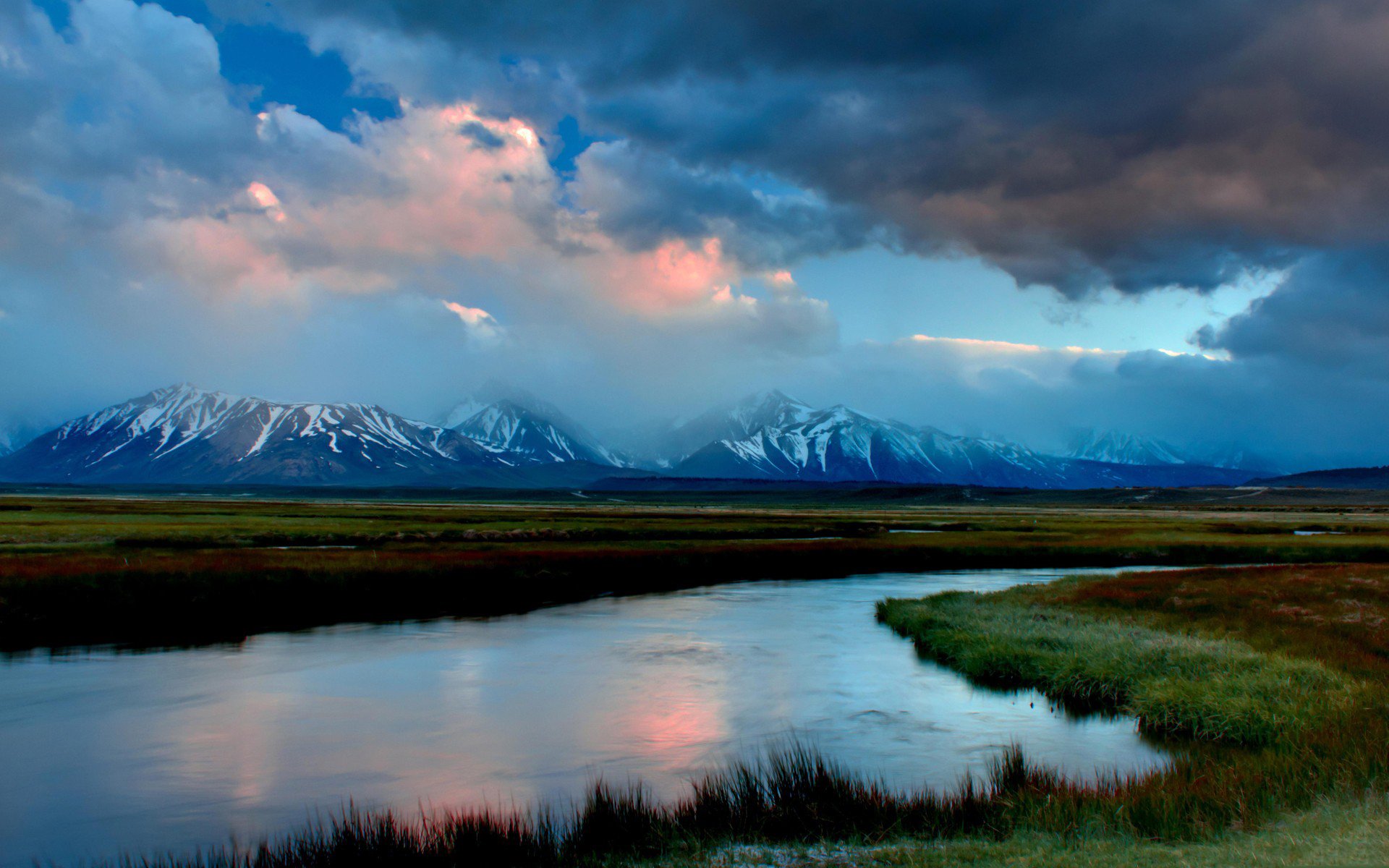 HD PC desktop wallpaper: nature landscape with a winding river reflecting pink-tinted clouds, grassy marshland and distant snow-capped mountains under a dramatic sky.