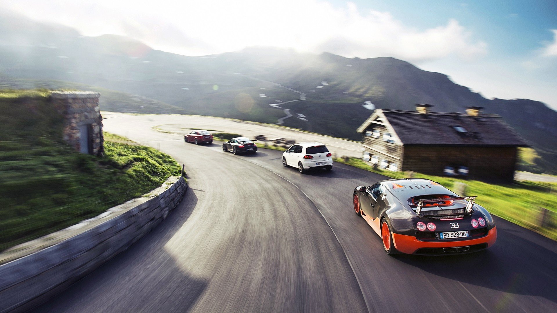 HD PC desktop wallpaper: an orange-and-black Bugatti leads a pack of sports vehicles along a winding mountain road past alpine houses under a dramatic sky.