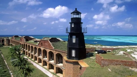 A stunning view of a black lighthouse overlooking turquoise waters in Florida, surrounded by historic man-made structures under a bright blue sky.