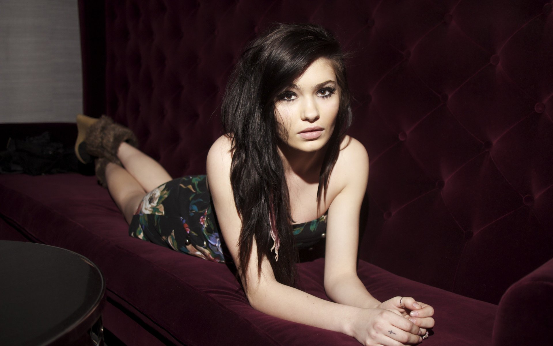 HD desktop wallpaper featuring Danielle Andersen, a woman with dark hair, lying on a deep burgundy velvet couch, gazing directly at the camera.
