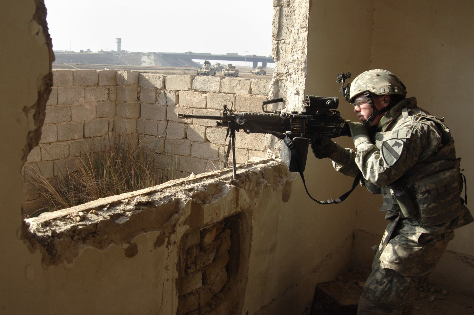HD PC desktop wallpaper of a soldier in military gear aiming a machine gun through a window in a partially damaged building.