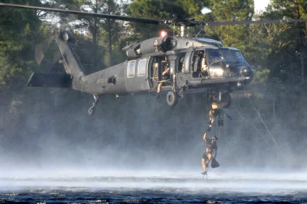 Military Sikorsky UH-60 Black Hawk helicopter hovers above water, deploying personnel during a tactical operation in a misty forest setting. HD PC desktop wallpaper.