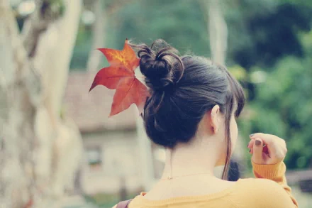 HD desktop wallpaper featuring the back of a woman with dark hair tied in a bun adorned with a red maple leaf, set against a softly blurred natural background.