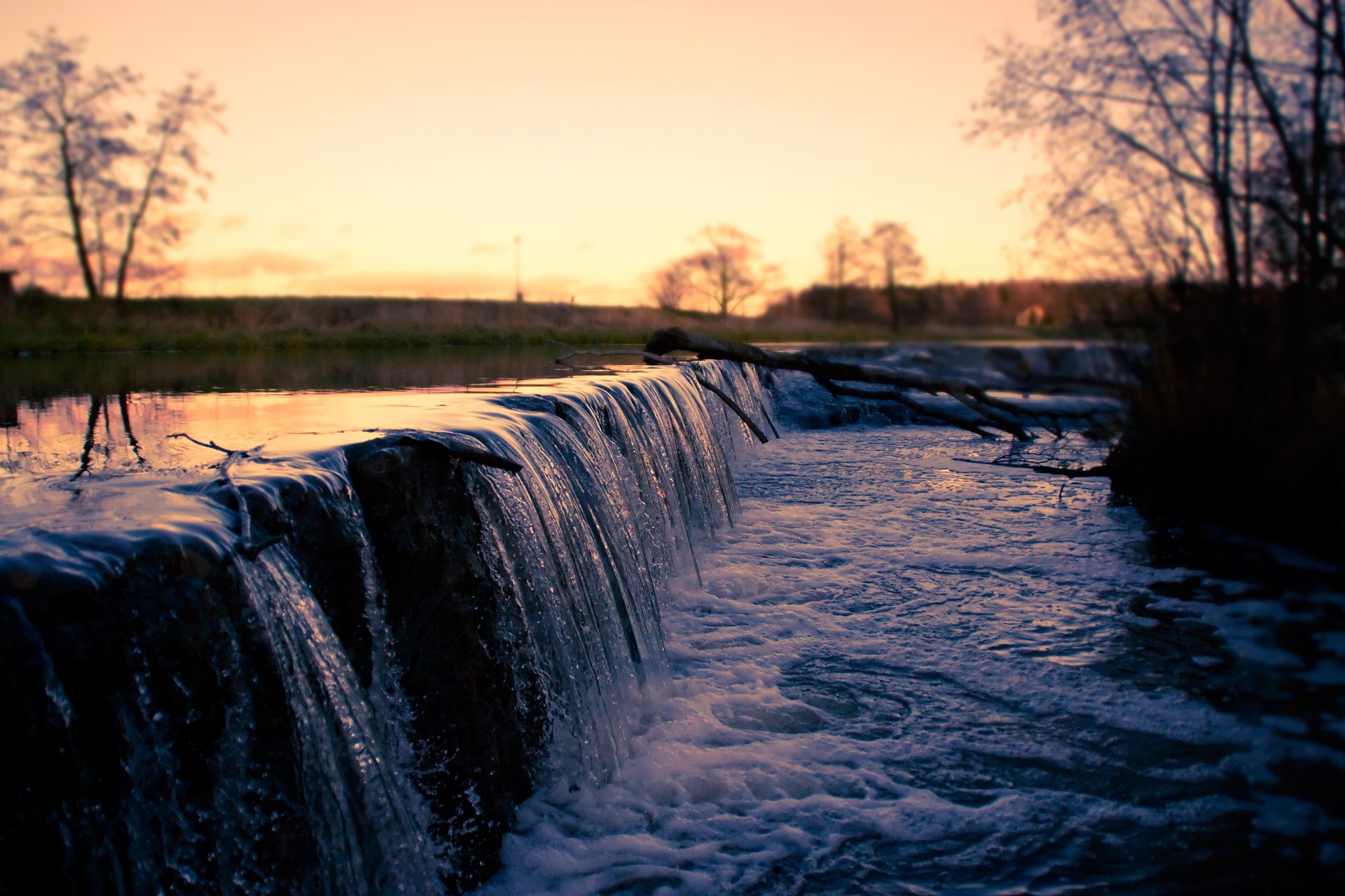 HD PC desktop wallpaper background: nature scene of a serene waterfall cascading over a low ledge at sunset, rippling river and silhouetted trees.