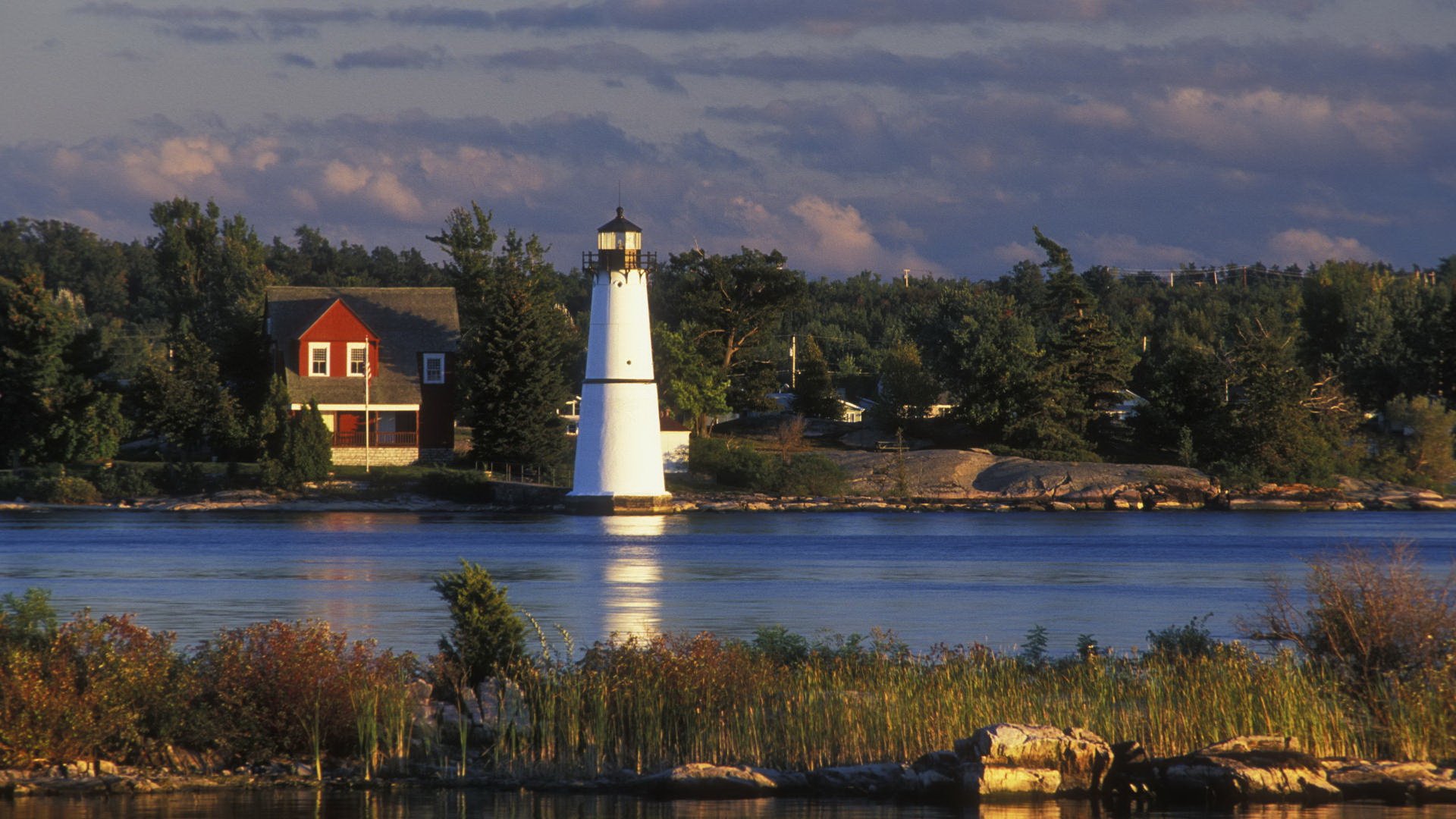 HD PC desktop wallpaper — serene lakeside with a man-made white lighthouse and red house on a tree-lined shore, reflected in calm water under a cloudy sky.