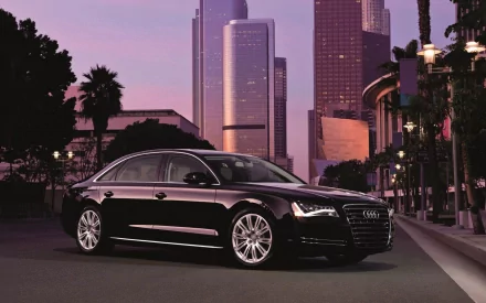 HD desktop wallpaper featuring a sleek black Audi A8 parked on a city street at dusk with tall buildings and palm trees in the background.