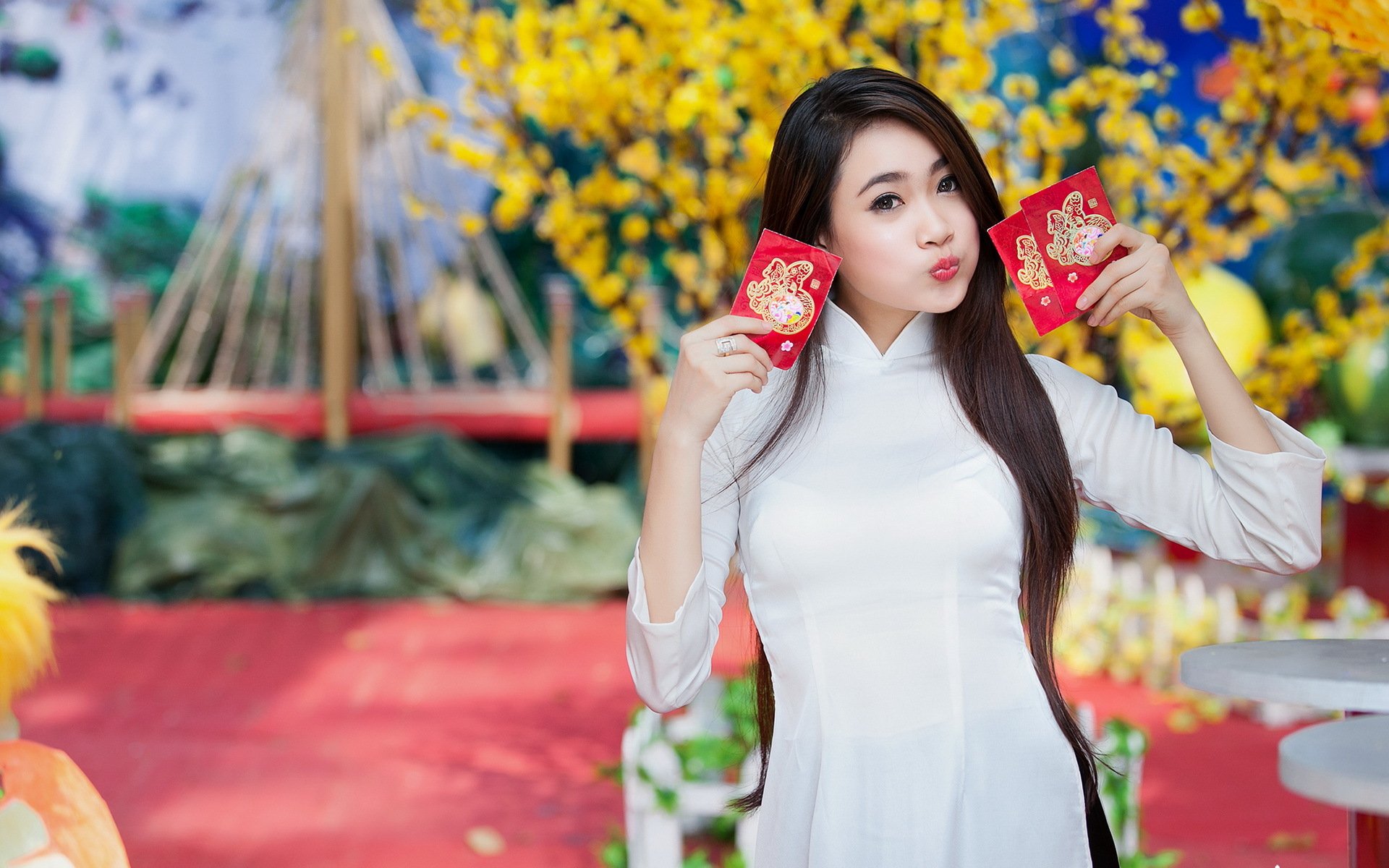 An Asian woman dressed in white holds two red envelopes, standing in front of a vibrant yellow tree and festive background. This HD desktop wallpaper captures a joyful moment.