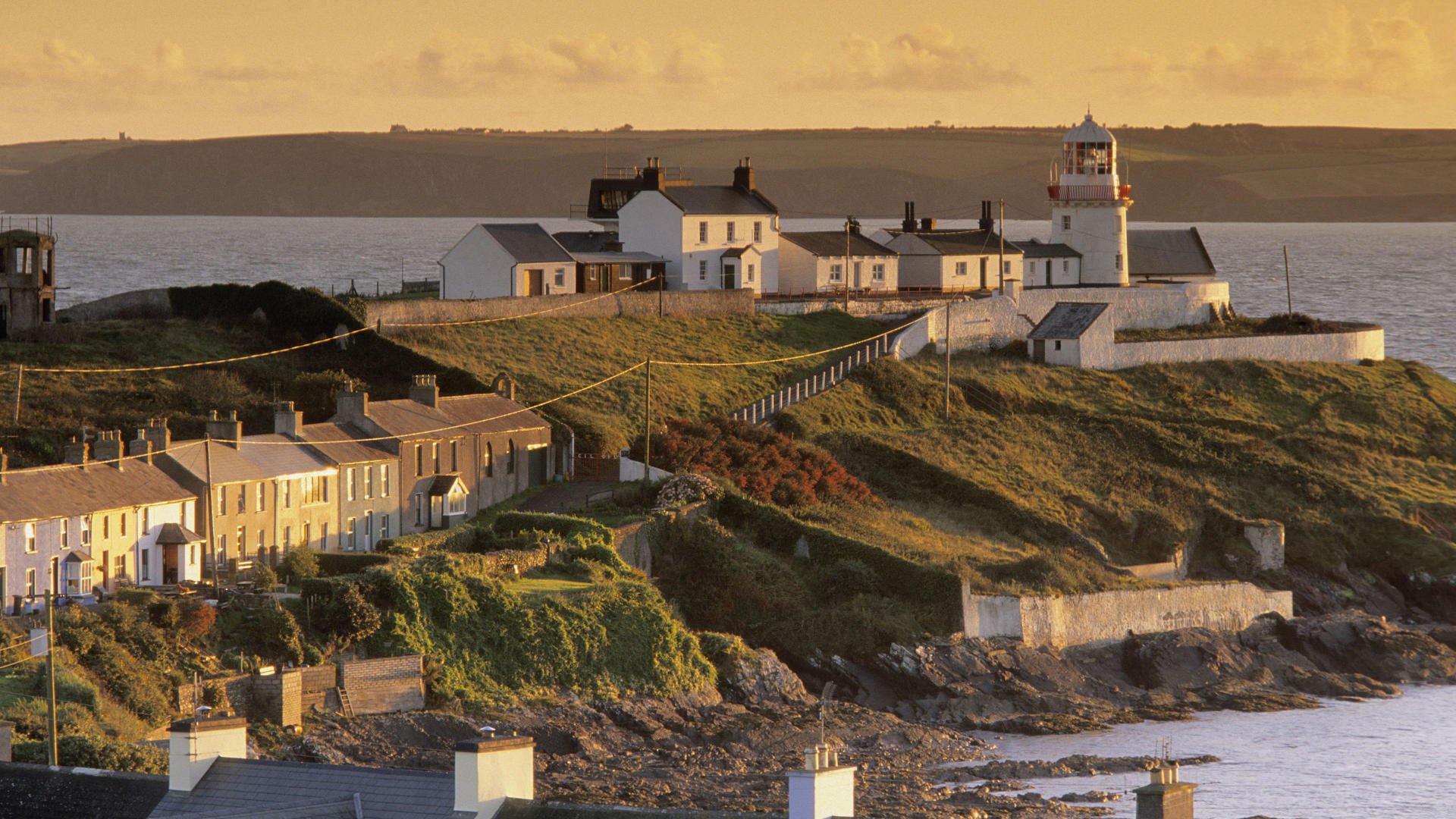 HD PC desktop wallpaper/background: man-made coastal village with a lighthouse on a sunlit headland, rows of houses and rocky shore.