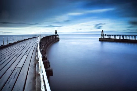 A serene HD desktop wallpaper showcasing a curving wooden pier extending into calm waters under a moody blue sky, highlighting man-made beauty in nature.