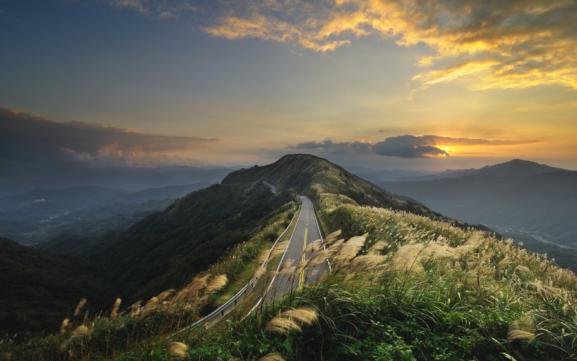 A serene HD wallpaper featuring a winding road traversing grassy hills beneath a colorful sunset, with majestic mountains in the background, creating a breathtaking natural landscape.