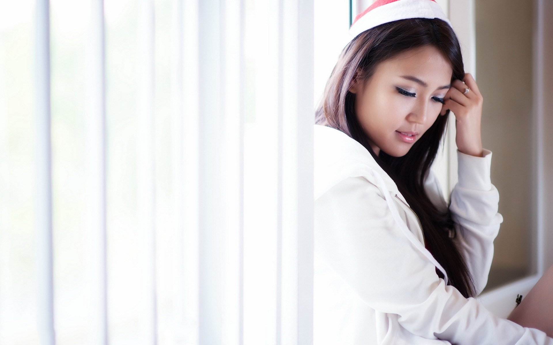 Asian woman in a Santa hat, sitting pensively by a window with white blinds. HD desktop wallpaper and background.