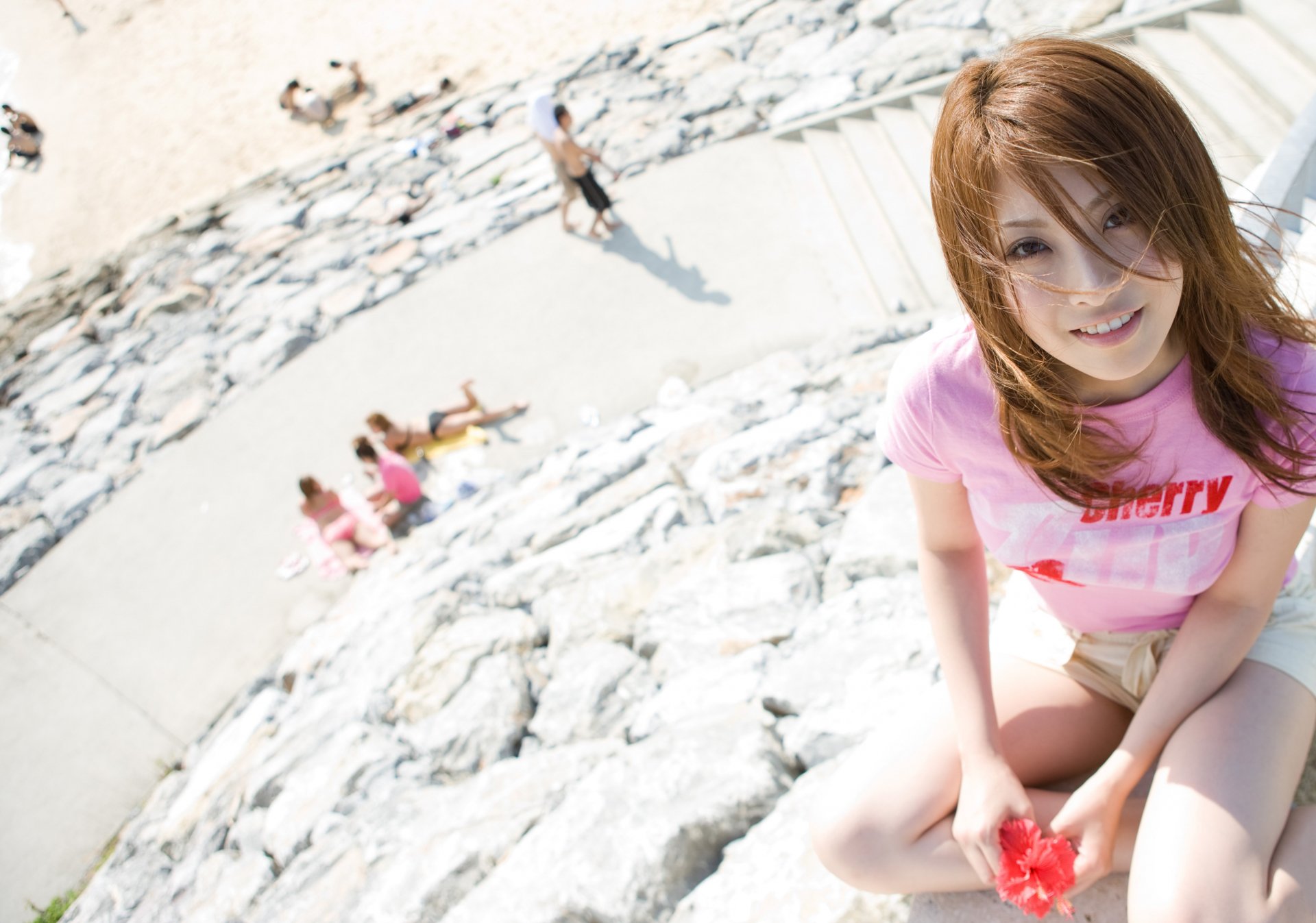 An Asian woman smiles while sitting on a rocky surface, with a beach scene in the background. This HD desktop wallpaper beautifully captures a moment of joy and relaxation.