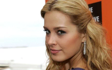 Close-up HD desktop wallpaper of a blonde female celebrity glancing over her shoulder, wearing dangling earrings and subtle makeup against a soft, neutral background.